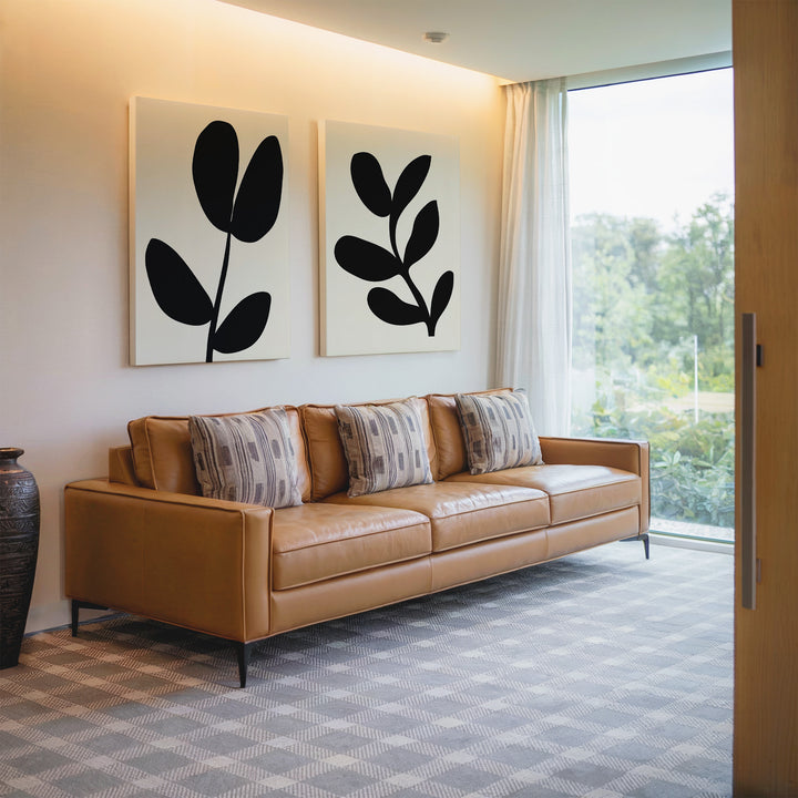 A modern living room with a tan leather sofa, four patterned cushions, the Black Leaf Set of 2 wall art above, a dark vase, and a large window overlooking greenery.