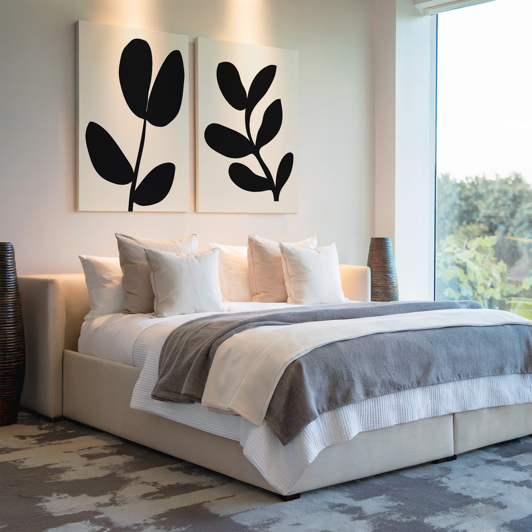 A modern bedroom with a large bed, white and gray bedding, multiple pillows, the Black Leaf Set of 2 wall art above the headboard, and a large window with a view of trees.