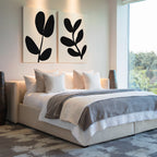 A modern bedroom with a large bed, white and gray bedding, multiple pillows, the Black Leaf Set of 2 wall art above the headboard, and a large window with a view of trees.