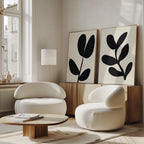 A modern, minimalist living room features two rounded white armchairs, a low wooden coffee table, and the Black Leaf Set of 2—large abstract botanical wall art displayed above a wood sideboard. Sunlight pours in through tall windows.