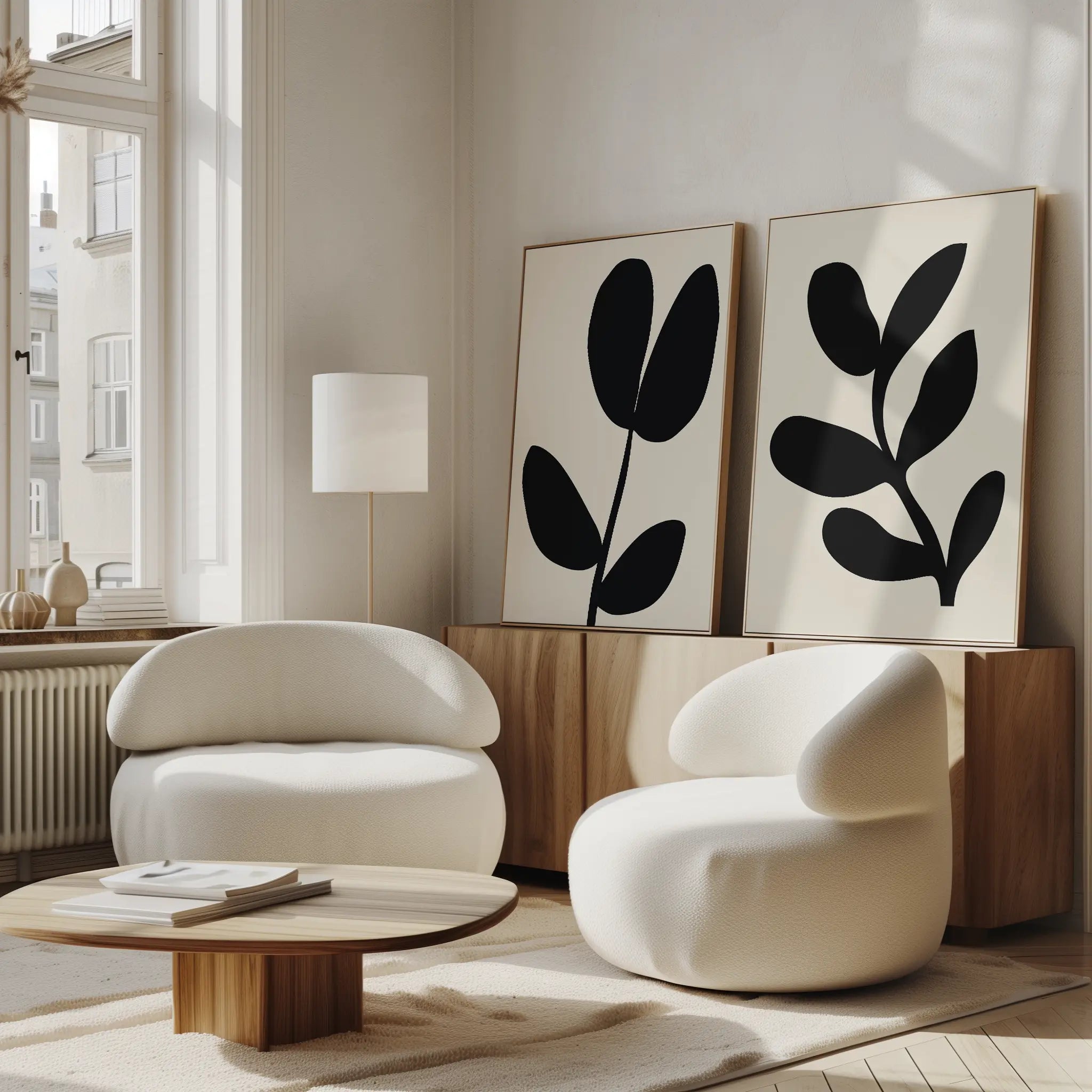 A modern, minimalist living room features two rounded white armchairs, a low wooden coffee table, and the Black Leaf Set of 2—large abstract botanical wall art displayed above a wood sideboard. Sunlight pours in through tall windows.