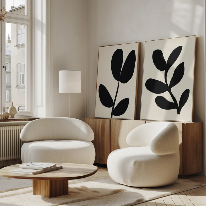A modern, minimalist living room features two rounded white armchairs, a low wooden coffee table, and the Black Leaf Set of 2—large abstract botanical wall art displayed above a wood sideboard. Sunlight pours in through tall windows.