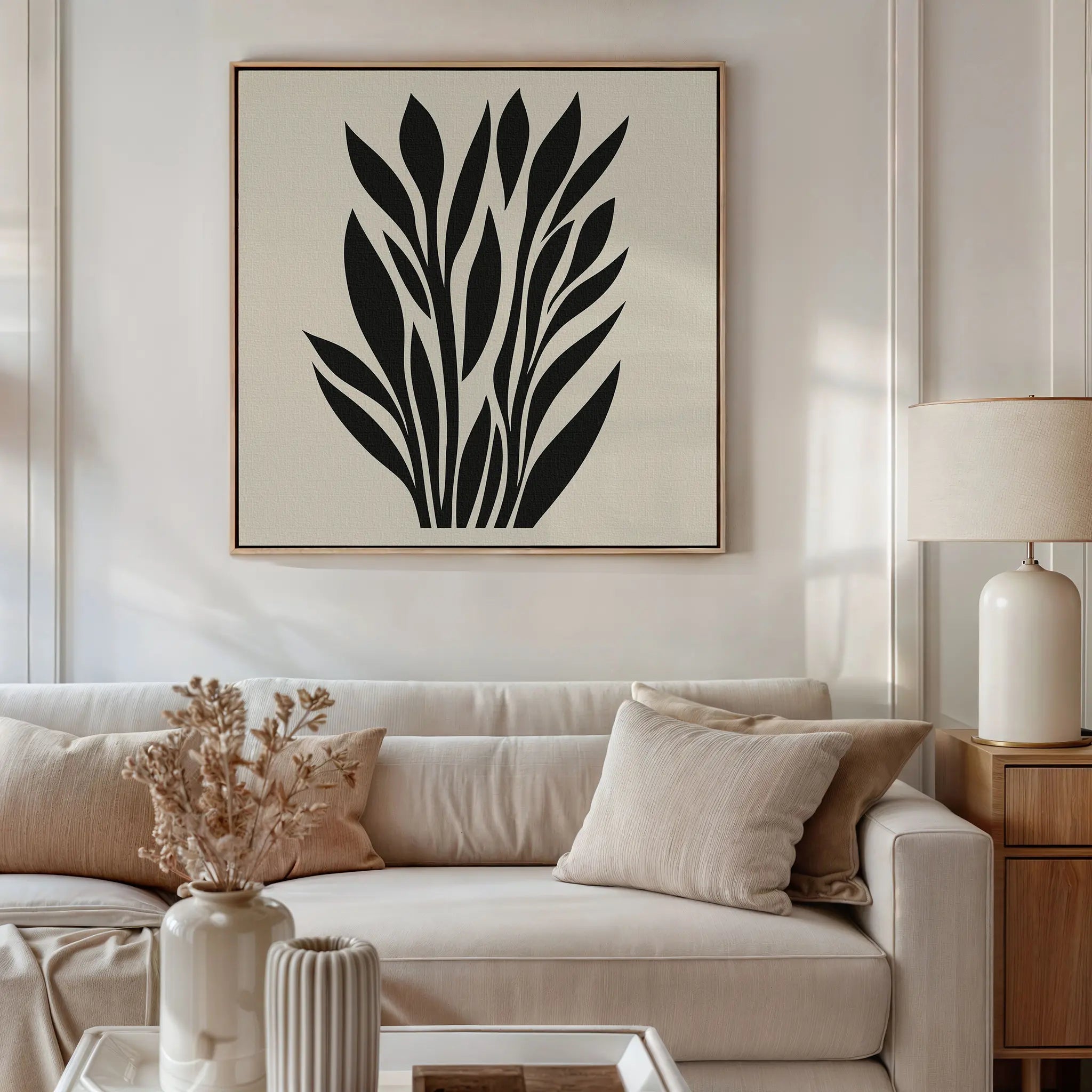 A modern living room with a beige sofa, neutral cushions, a wooden side table and white lamp, minimalist decor, and the Black Palm Study Canvas Art with abstract black palm leaves displayed above the sofa.