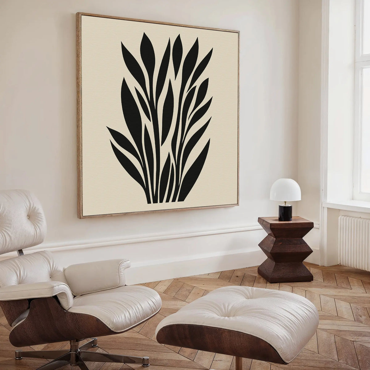 A modern living room features a cream lounge chair and ottoman, a side table with a white lamp, and the Black Palm Study Canvas Art. Light wood herringbone floors and large windows complete this stylish interior.