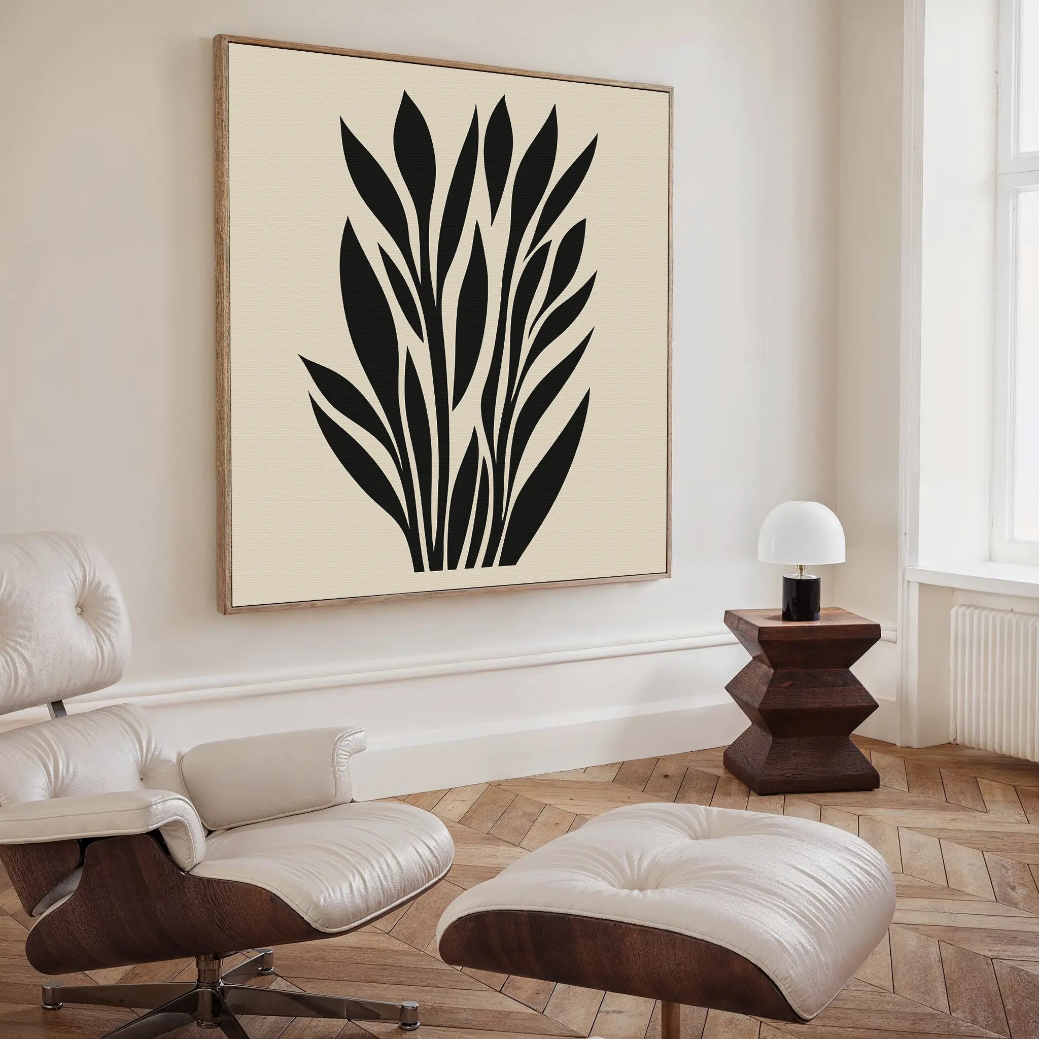 A modern living room features a cream lounge chair and ottoman, a side table with a white lamp, and the Black Palm Study Canvas Art. Light wood herringbone floors and large windows complete this stylish interior.