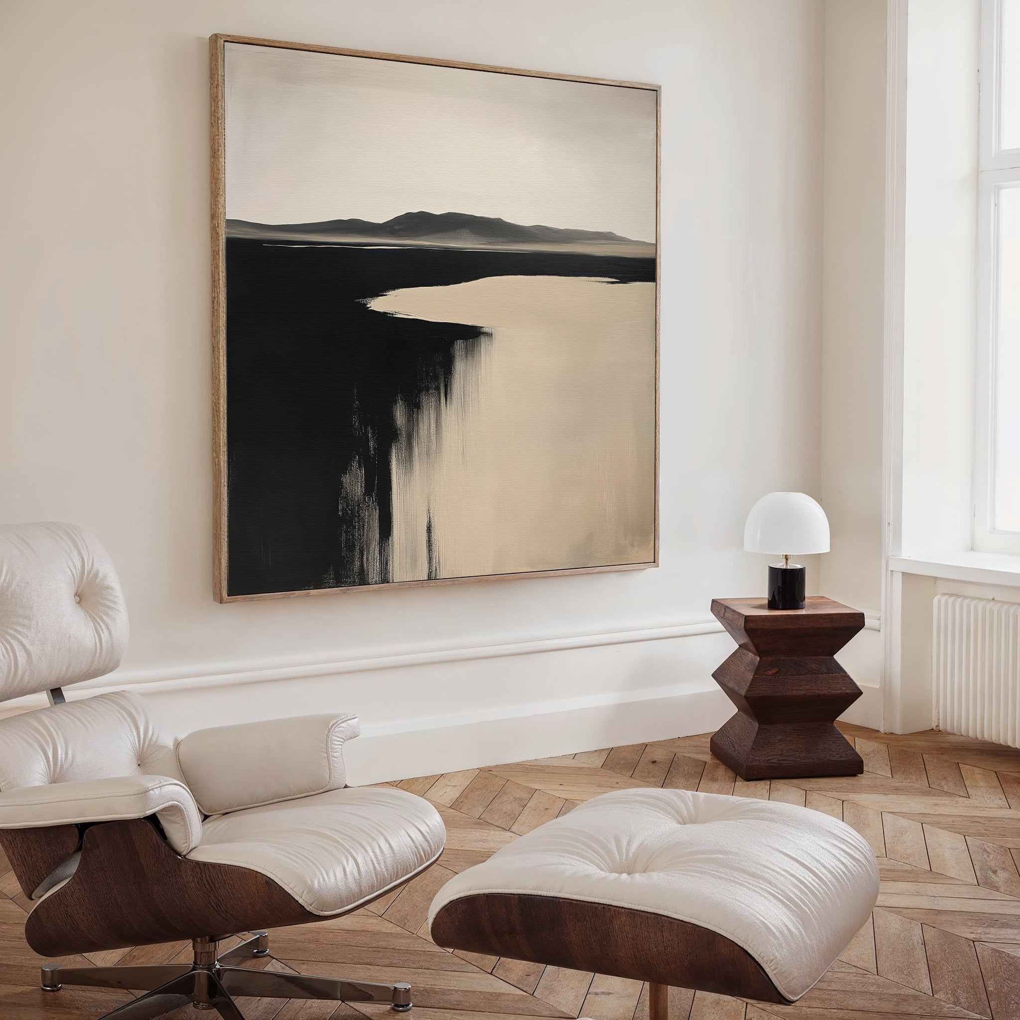 A modern, minimalist room with a white leather lounge chair and ottoman, the Black Shore Canvas Art above, a geometric wooden side table, and a small white lamp by a large window on chevron-patterned wood floors.
