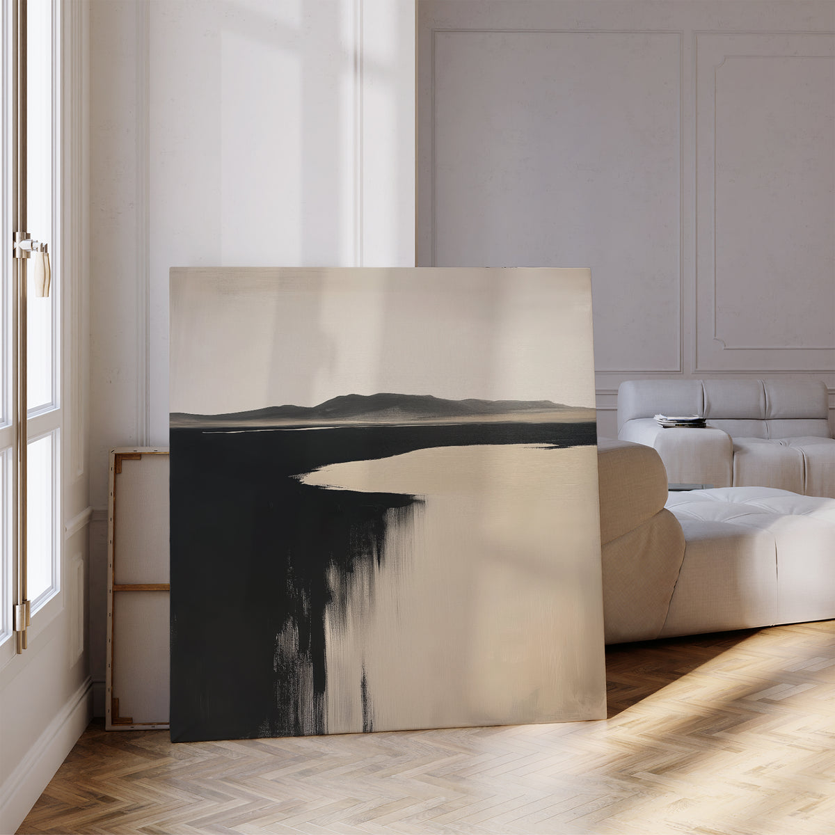 The Black Shore Canvas Art, featuring dark and light tones, leans against a wall in a bright modern living room with white walls, parquet floors, a beige sofa, and sunlight pouring through tall windows.