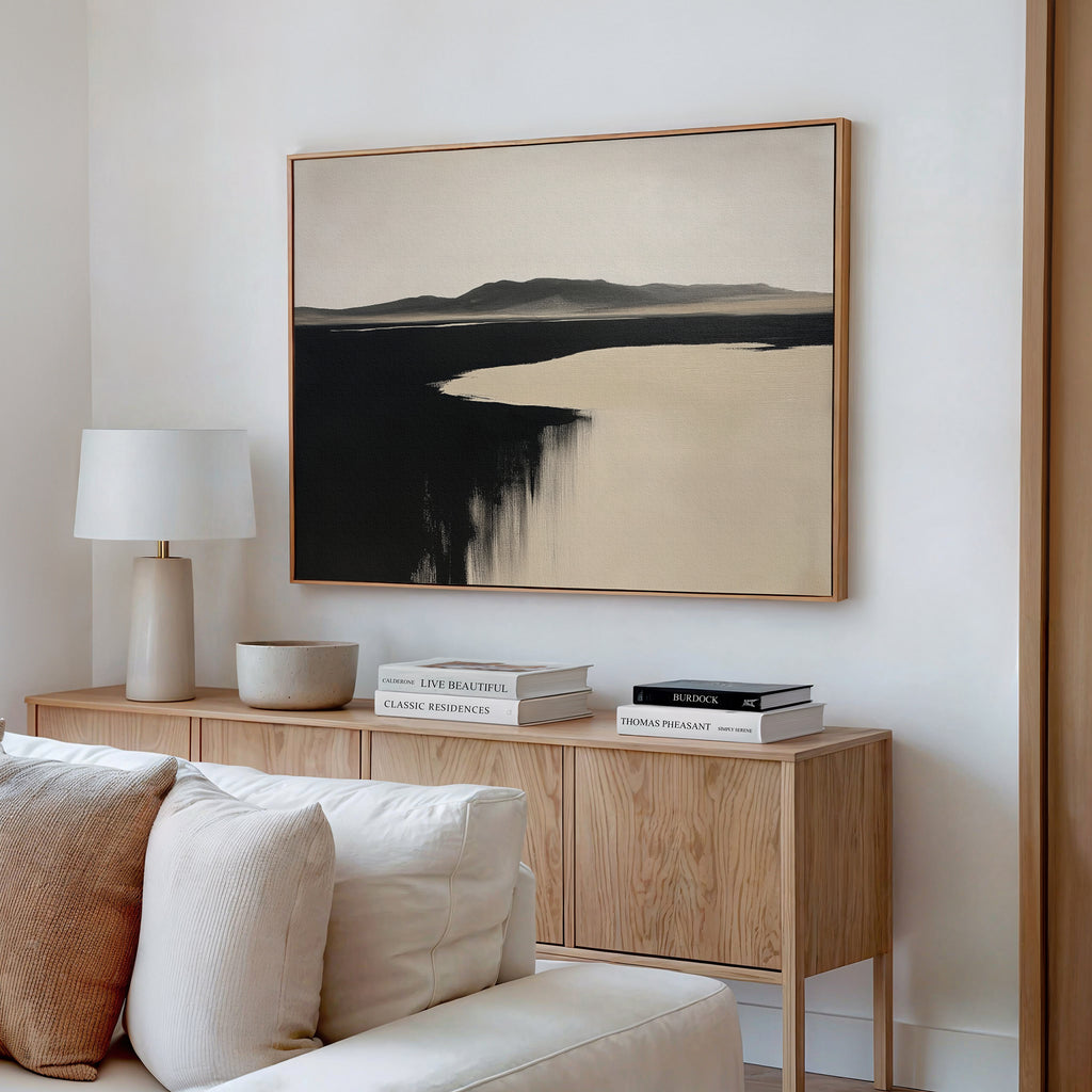 A minimal living room with a light wood console, stacked books, white lamp, bowl, and the Black Shore Canvas Art – Horizontal in neutral tones above a white sofa—ideal for fans of modern living room decor.