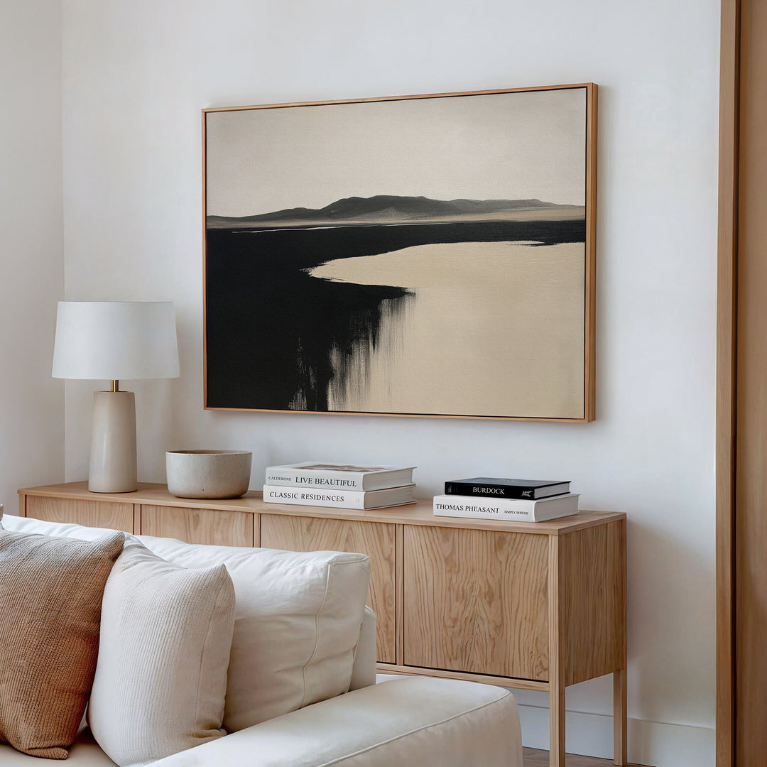A minimal living room with a light wood console, stacked books, white lamp, bowl, and the Black Shore Canvas Art – Horizontal in neutral tones above a white sofa—ideal for fans of modern living room decor.