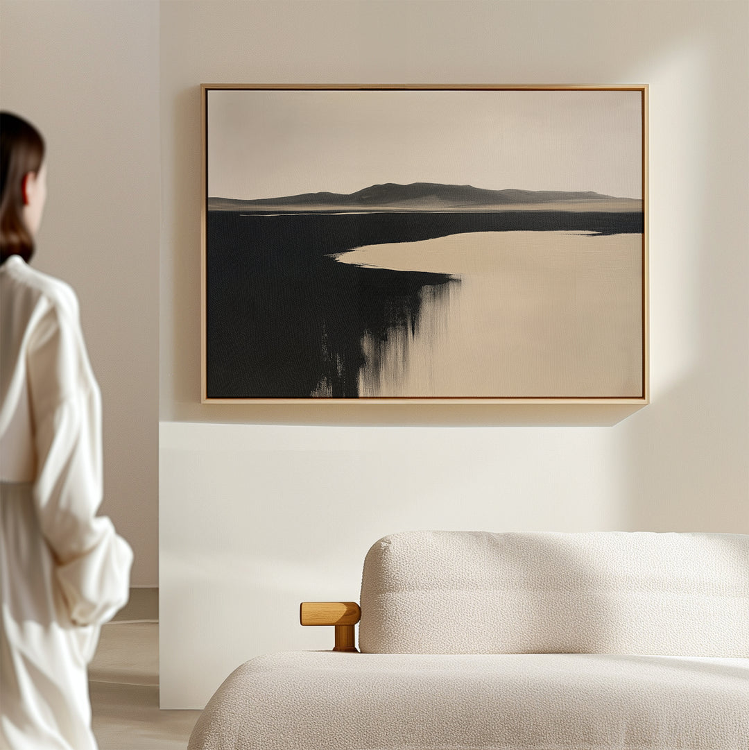 A person stands in a minimalist room, admiring the Black Shore Canvas Art – Horizontal with abstract dark and light tones above a light sofa, adding elegant style to the modern living room decor.