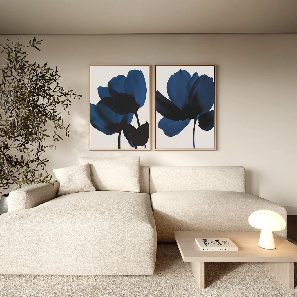 A modern living room with a light beige sectional, a small coffee table with books and a glowing lamp, a tall plant, and the Bloom Set of 2 blue contemporary wall art prints inspired by Nordic Park.