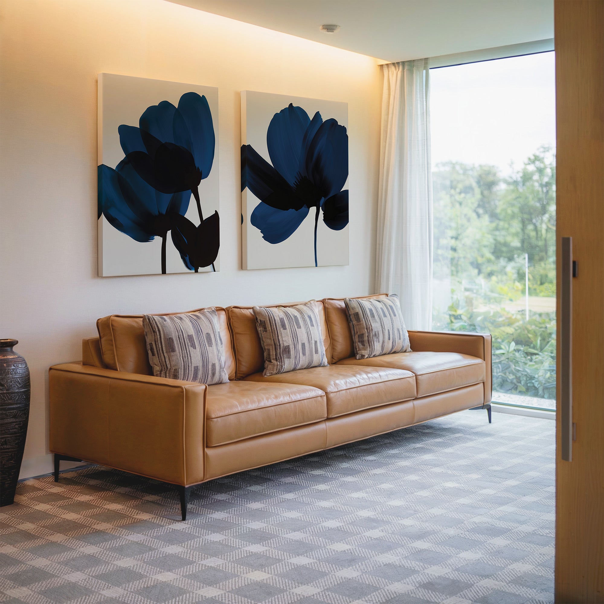 A modern living room featuring a tan leather sofa with patterned cushions, Bloom Set of 2 blue floral wall art, a floor vase, and a large window with sheer curtains overlooking Nordic Parks lush greenery.