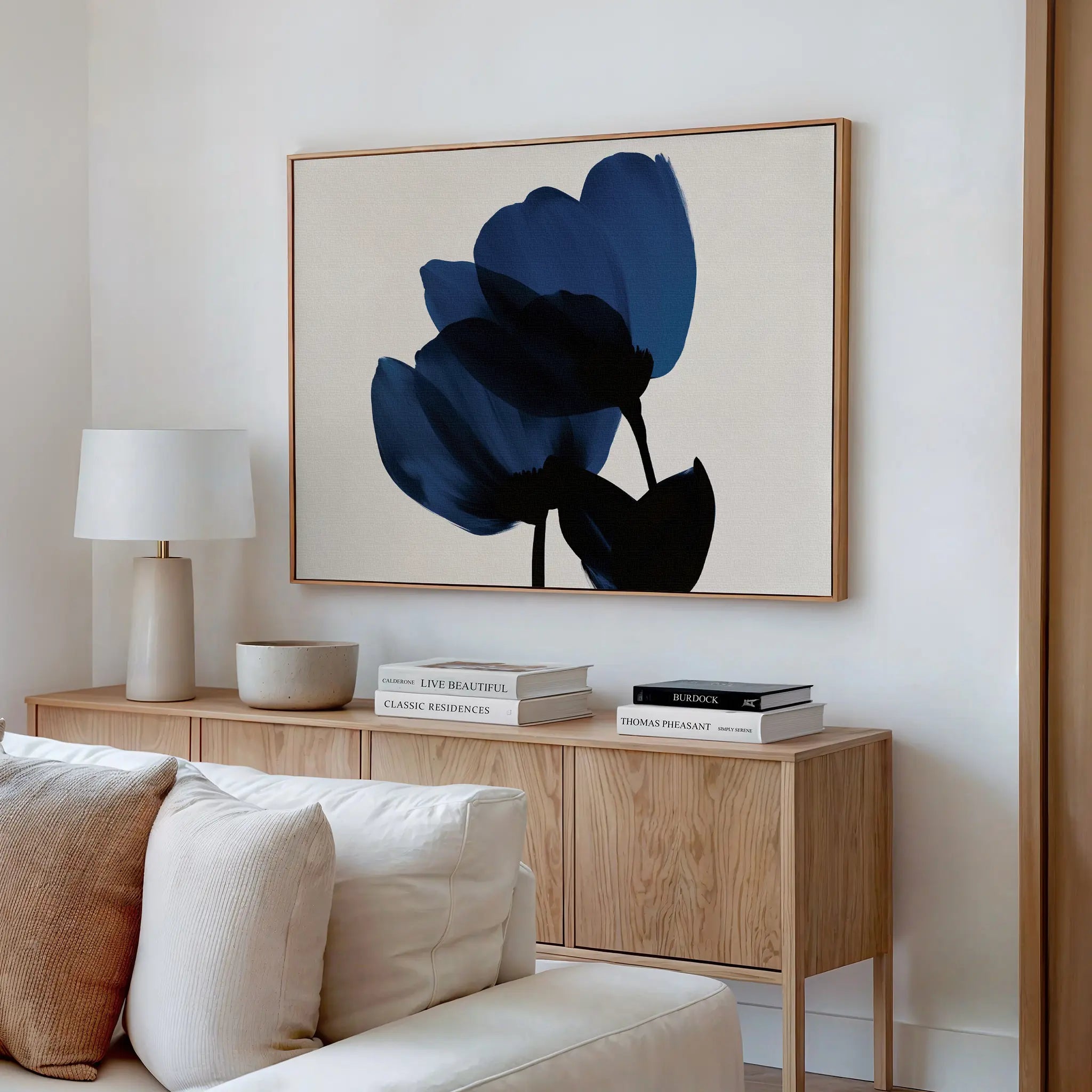 A minimalist living room features a light wood sideboard with books and decor, a table lamp, and the Bloom Abstract Canvas Art – Horizontal, a large framed blue floral painting, displayed above a cream-colored sofa.