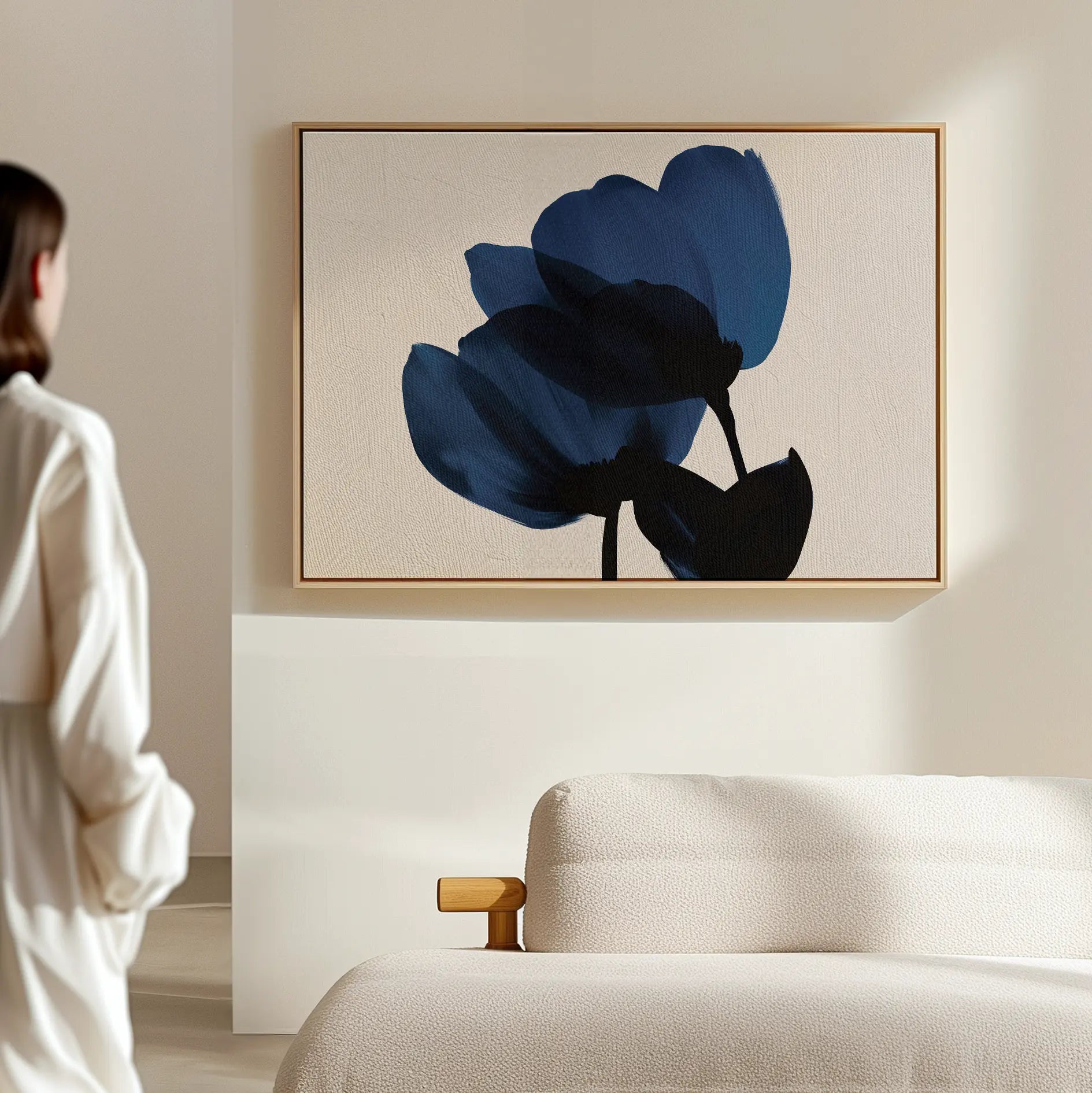 A person in a white robe stands near a modern beige sofa, admiring the Bloom Abstract Canvas Art – Horizontal, which features two dark blue flowers on a light textured wall—a perfect addition to contemporary living room decor.