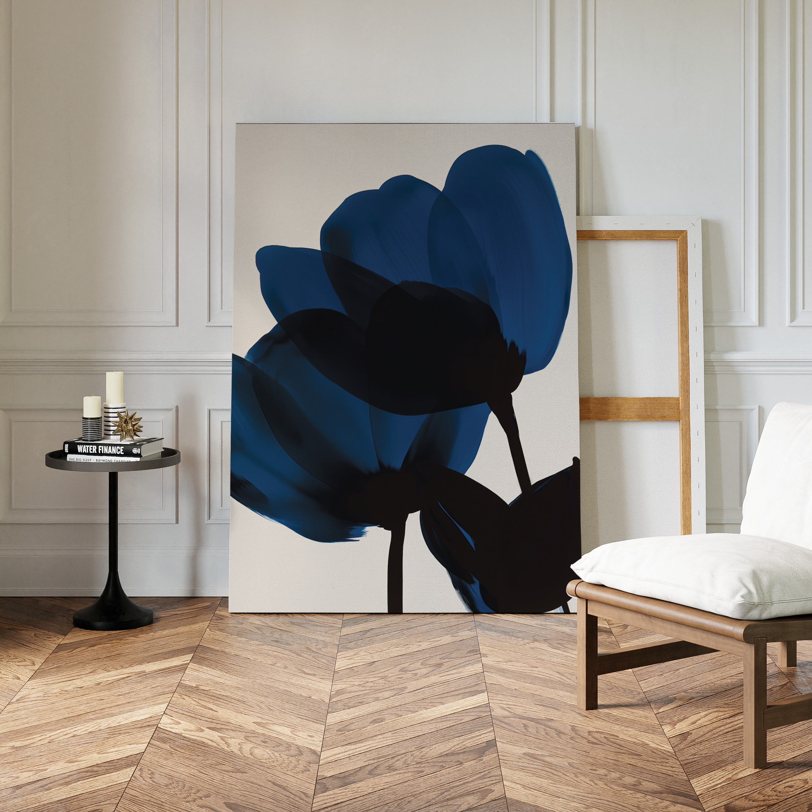 The Bloom Abstract Canvas Art, featuring bold blue and black flower shapes, leans against a white paneled wall beside an empty frame. Candles on a small table and a cushioned chair complete the stylish living room decor.