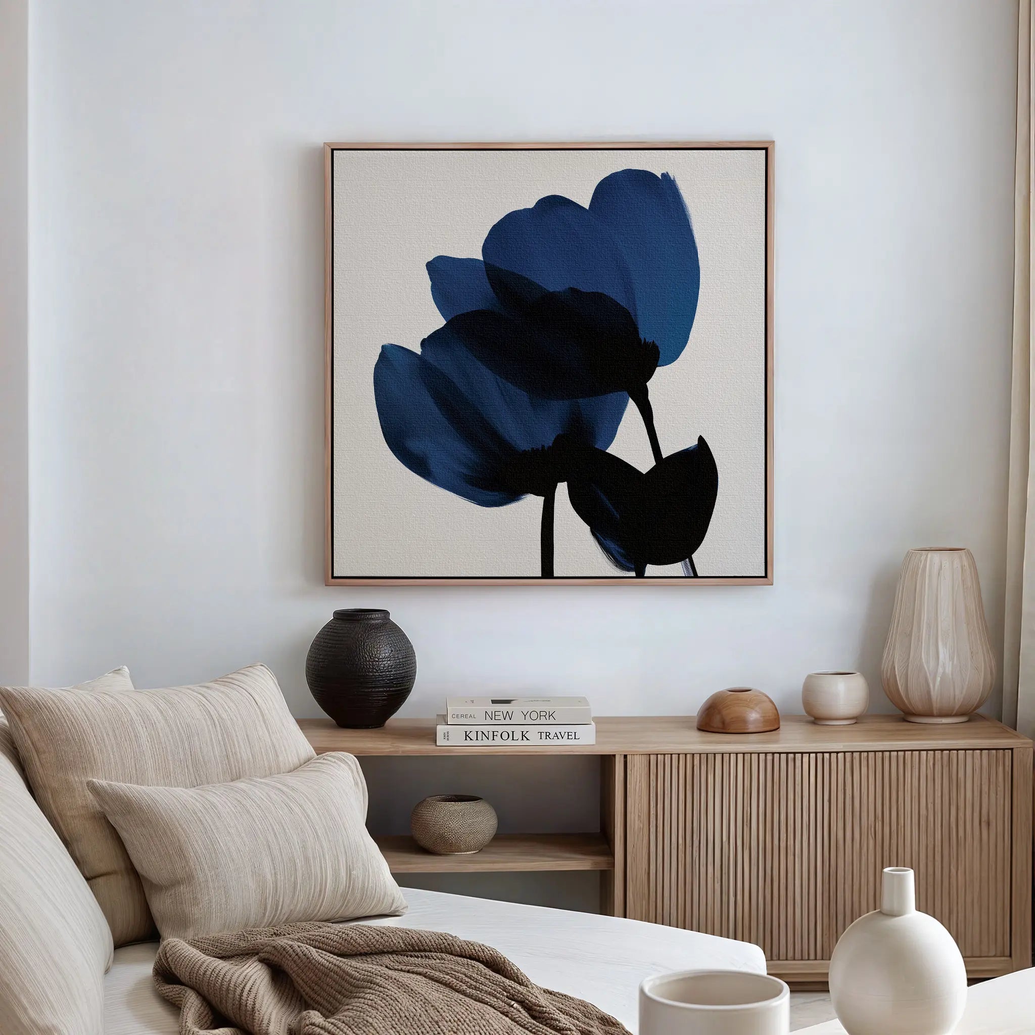 A modern living room with a beige sofa, neutral cushions, wooden sideboard, decorative vases, books, and the Bloom Abstract Canvas Art – Square featuring abstract blue flowers on a white wall.