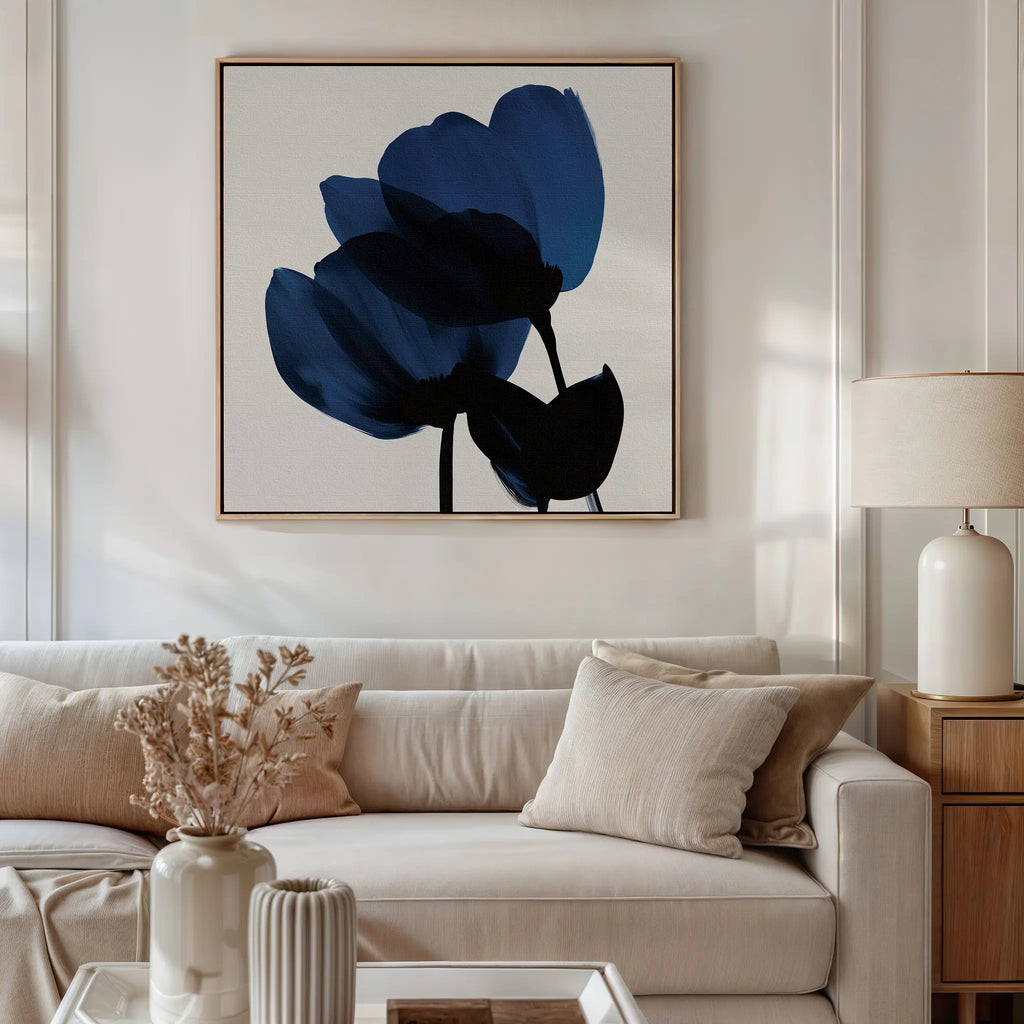 A modern living room features a white sofa, neutral pillows, a table lamp, and dried flowers. Above the sofa hangs the Bloom Abstract Canvas Art – Square, showcasing abstract blue flowers that add style to the minimalist decor.
