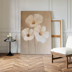 The Blossom Trio Abstract Canvas Art, a large minimalist piece with three white flowers, leans against a stylish rooms wall with herringbone floors, beside an empty frame, white cushioned chair, and small black table with candles.