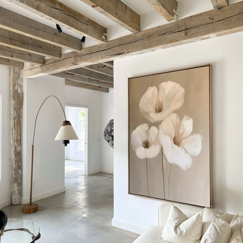 The Blossom Trio Abstract Canvas Art, featuring three white flowers, hangs on a white wall in a bright modern living room with exposed wooden ceiling beams, a floor lamp, and minimalist decor.
