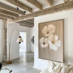 The Blossom Trio Abstract Canvas Art, featuring three white flowers, hangs on a white wall in a bright modern living room with exposed wooden ceiling beams, a floor lamp, and minimalist decor.