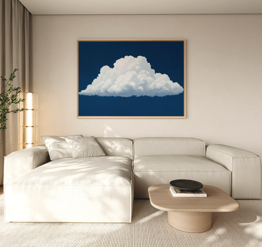 A modern living room with a beige sectional sofa, light wood coffee table, plant, and the Blue Horizon Cloud Canvas Art—a minimalist piece depicting a fluffy white cloud on a deep blue background—hanging above.
