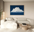 A modern living room with a beige sectional sofa, light wood coffee table, plant, and the Blue Horizon Cloud Canvas Art—a minimalist piece depicting a fluffy white cloud on a deep blue background—hanging above.
