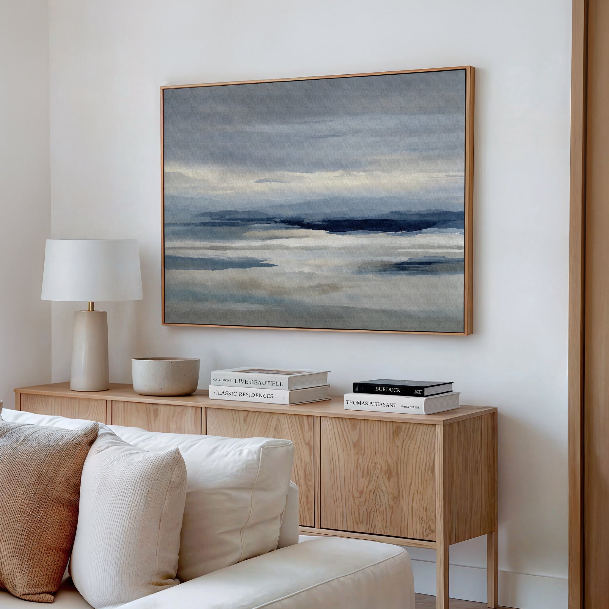 A modern living room features a light wood sideboard with stacked books, a white lamp, and the Blue Mist Horizon Canvas Art above, adding minimalist style with its blue and gray tones.