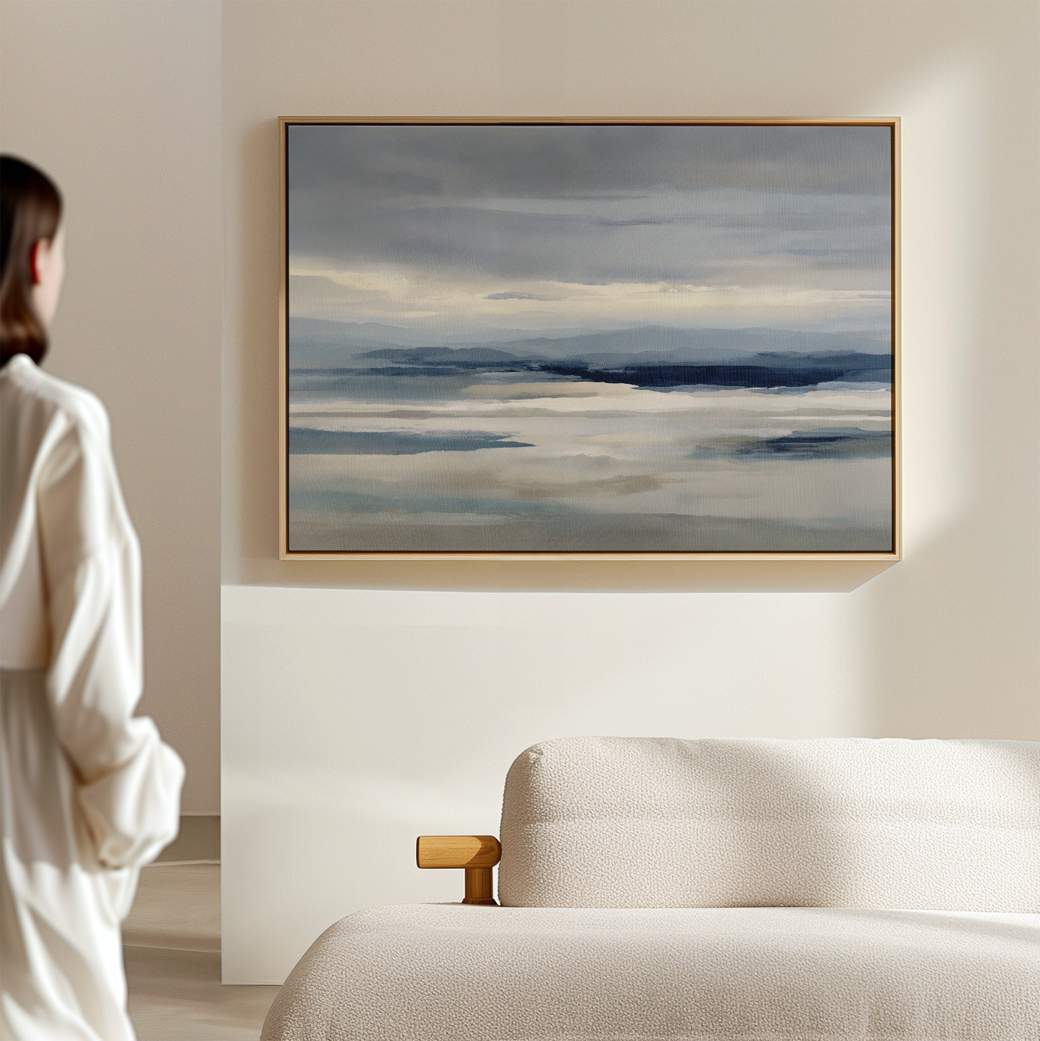 A person stands in a minimal, light-filled room admiring the Blue Mist Horizon Canvas Art with muted blue and gray tones above a cream sofa, adding an elegant focal point to modern living room decor.