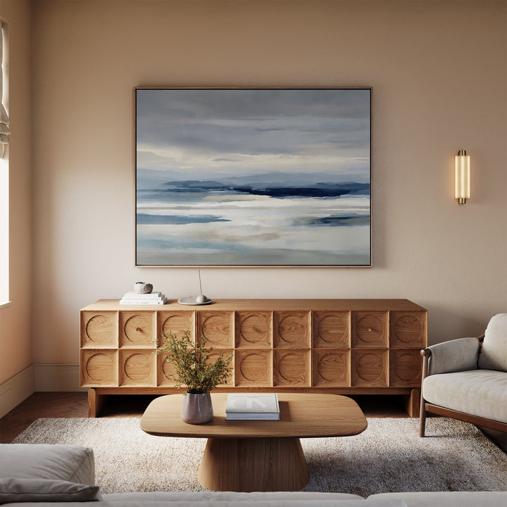A minimalist living room features a beige sofa, wooden coffee table, textured sideboard, and the Blue Mist Horizon Canvas Art in blue and white above. A modern wall sconce and a vase with flowers complete the decor.