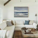 A cozy living room with white shiplap walls, a large white sectional sofa with assorted throw pillows, a wooden coffee table, and Blue Mist Horizon Canvas Art above the sofa completes the inviting decor.
