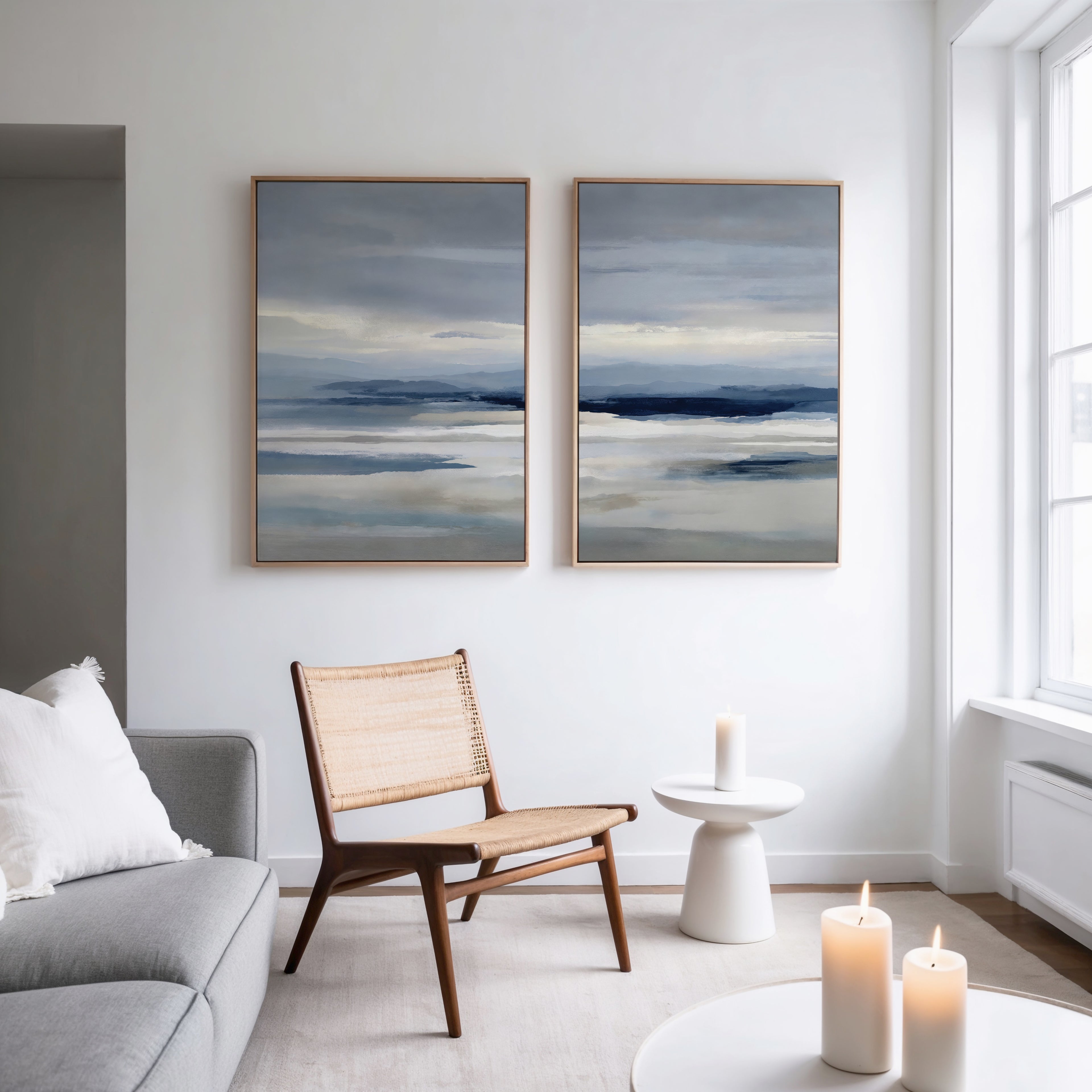 A modern, minimalist living room featuring a light sofa, wooden chair, two lit candles, a small round table, and the Blue Mist Horizon Set of 2—striking blue and gray minimalist canvas wall art for refined decor.