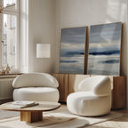 A modern living room features two rounded white chairs, a low wood coffee table, and a light wood sideboard topped with the Blue Mist Horizon Set of 2 canvas prints in blue and gray. Sunlight streams through tall windows, brightening the decor.