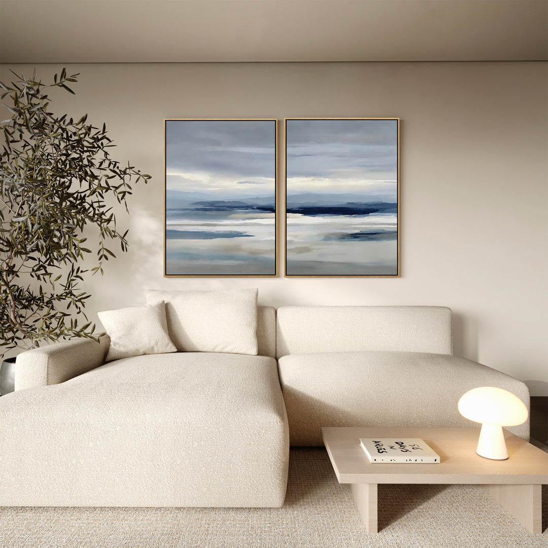 A modern living room featuring a cream sectional, small wooden table with lamp and book, leafy plant, and minimalist wall art—Blue Mist Horizon Set of 2 abstract paintings—adds the perfect touch of living room decor.