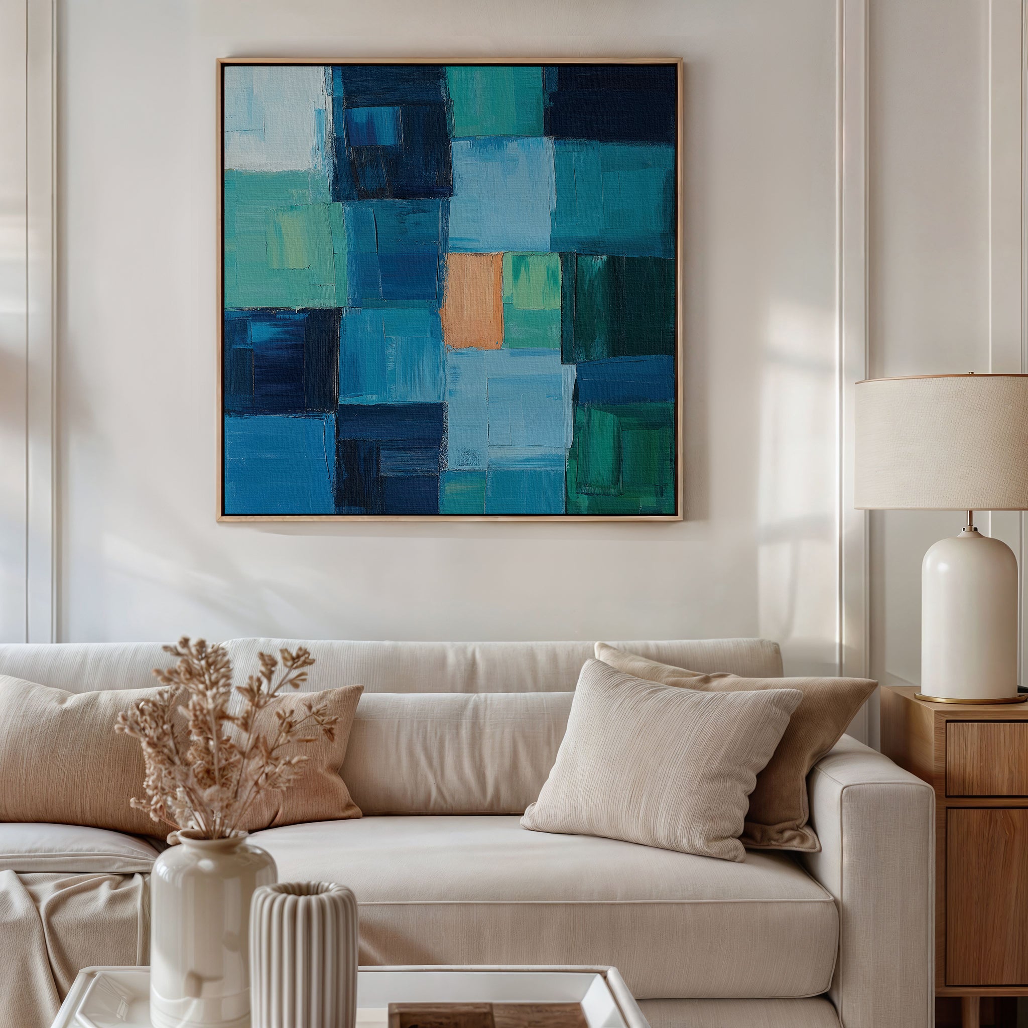 A modern living room with a beige sofa, light cushions, a side table with a white lamp, a vase of dried flowers, and Blue Mosaic Abstract Canvas Art featuring blue and green squares as the focal point of the decor.