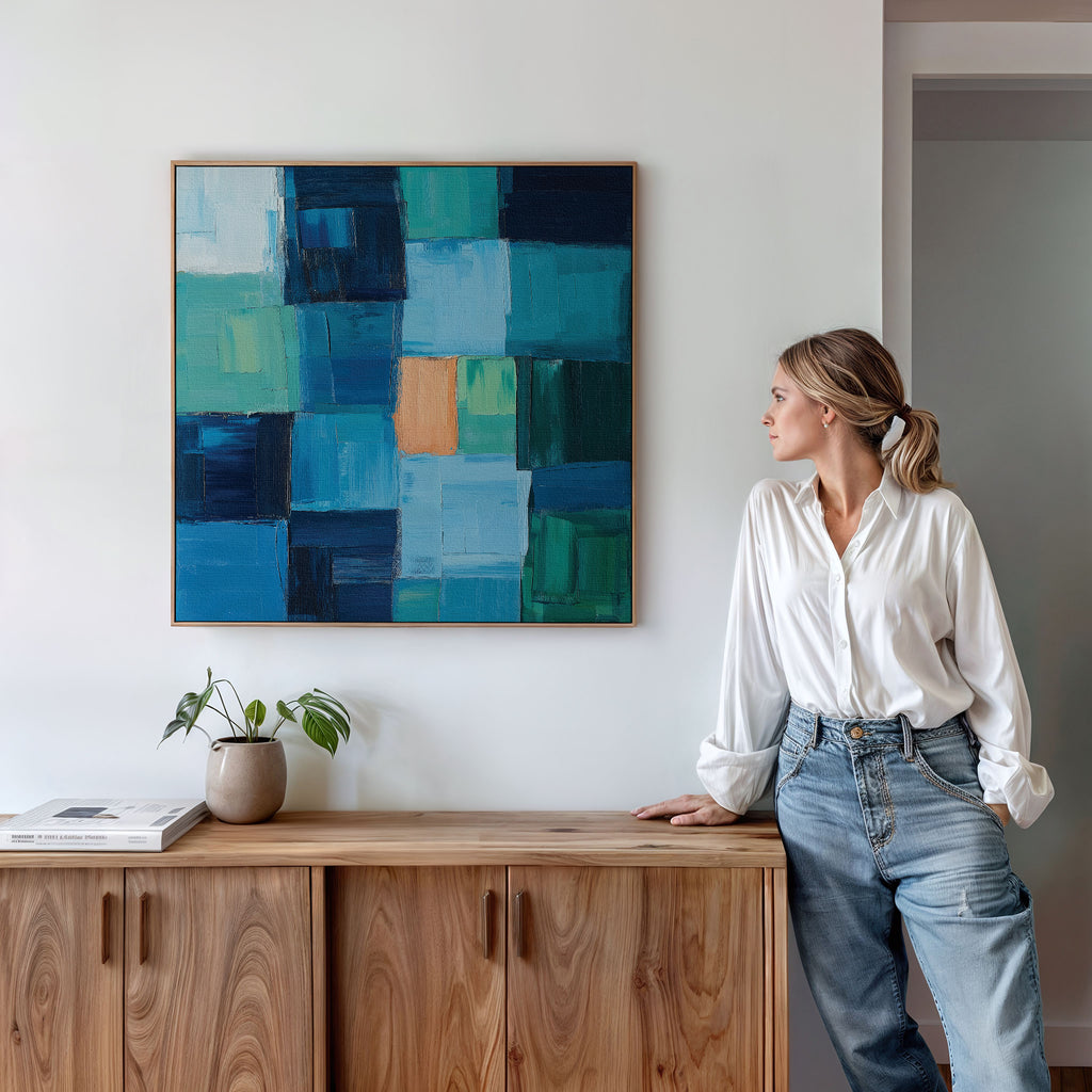 A woman in a white shirt and jeans leans on a wooden cabinet, admiring Blue Mosaic Abstract Canvas Art with blue and green squares. Perfect for living room decor, the scene also features a small plant and book on the cabinet against a white wall.