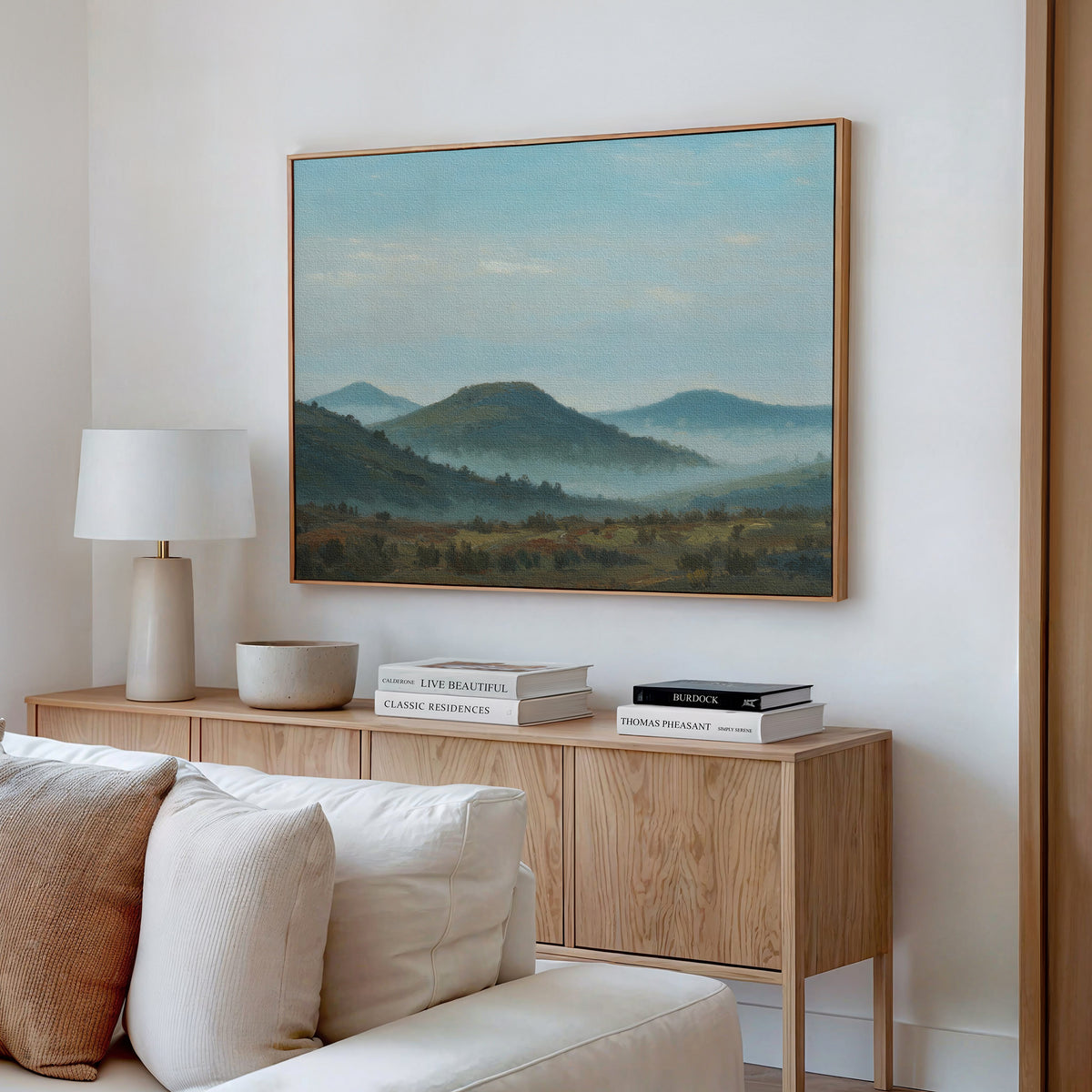 A modern living room features a beige sofa, wooden sideboard, table lamp, and stacked books. Above the sideboard, the Blue Ridge View Canvas Art with misty green hills adds a serene touch to the white wall.