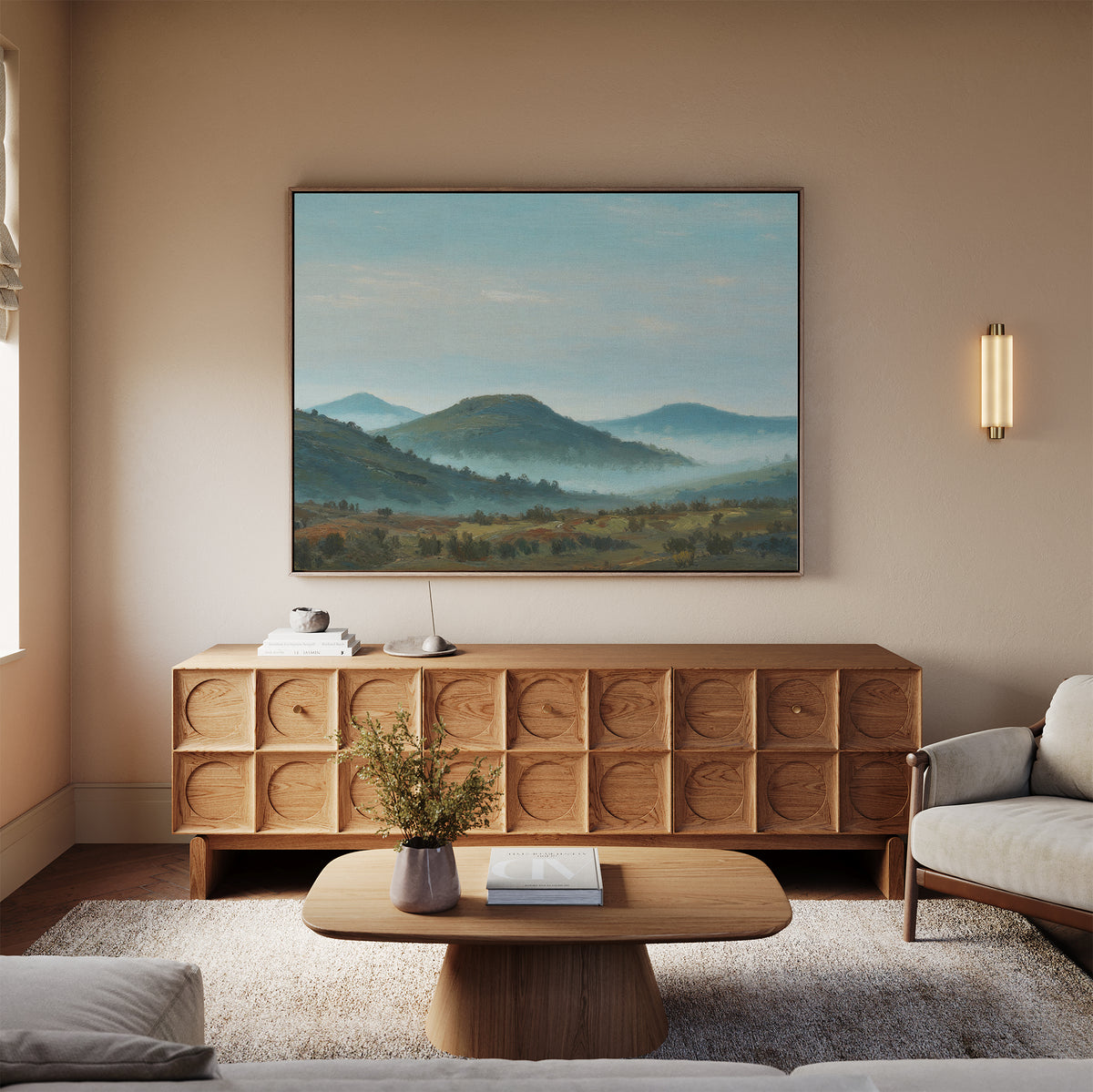 A neutral-toned living room with a wooden sideboard, a coffee table with a vase, a beige sofa, the Blue Ridge View Canvas Art of mountains on the wall, and a modern sconce light.