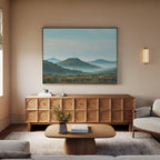 A neutral-toned living room with a wooden sideboard, a coffee table with a vase, a beige sofa, the Blue Ridge View Canvas Art of mountains on the wall, and a modern sconce light.