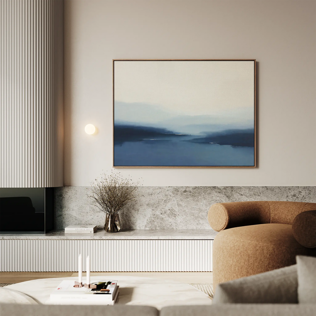 A modern living room features the Blue Valley Mist Canvas Art above a marble shelf, a curved brown sofa, a round light fixture, and minimalist decor like a vase with dried branches on the shelf.