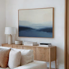 A modern living room features the Blue Valley Mist Canvas Art in blue and beige tones above a light wood sideboard with a white lamp, decorative bowl, and stacked books. A cream sofa sits in front for a minimalist decor look.