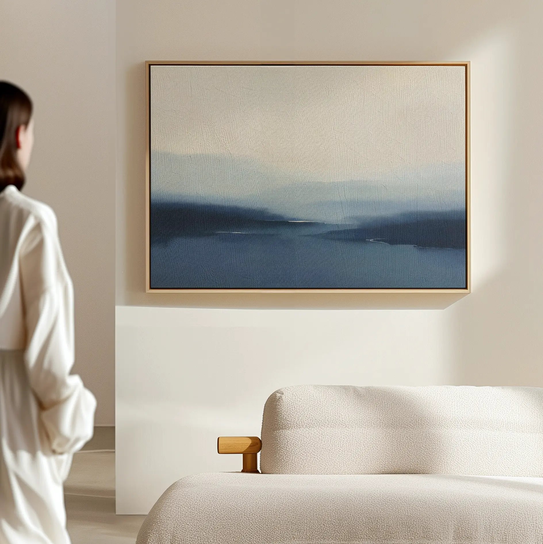 A person in white stands by a beige sofa, admiring the Blue Valley Mist Canvas Art—a large landscape print in blue and cream tones that enhances the minimalist decor of the modern room.