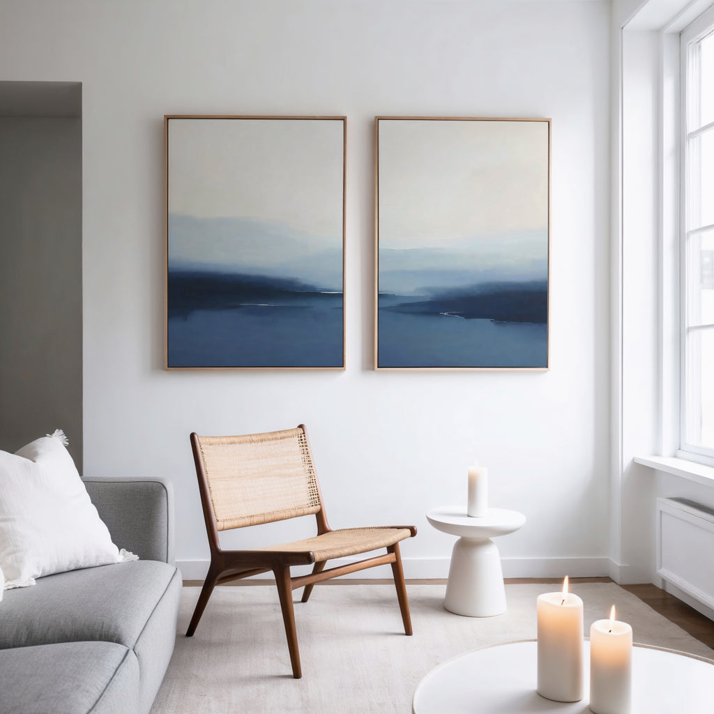 A bright, minimalist living room features a wooden chair, a white side table with a lit candle, a gray sofa, and the Blue Valley Mist Set of 2 wall art prints. Two more lit candles glow softly on a table in the foreground.