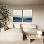 A modern living room with a cream sectional, small wood coffee table with lamp and book, minimalist decor, and the Blue Valley Mist Set of 2 wall art prints above the sofa. A tall plant stands in the corner.