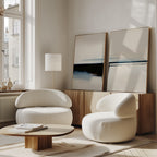 A modern living room features two rounded white armchairs, a low wooden coffee table, and the Blurred Reflections Set of 2 minimalist wall art on a wooden sideboard. Large windows flood the cozy space with natural light.