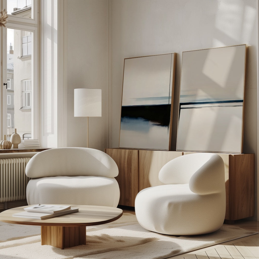 A modern living room features two rounded white armchairs, a low wooden coffee table, and the Blurred Reflections Set of 2 minimalist wall art on a wooden sideboard. Large windows flood the cozy space with natural light.