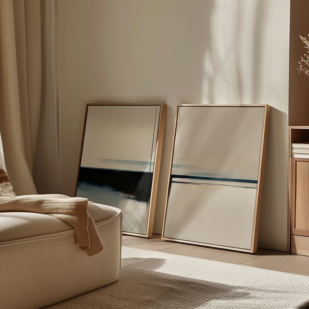 The Blurred Reflections Set of 2 features abstract framed art with horizontal dark and blue lines, leaning against a beige wall in a bright living space, paired with a cozy cream chair for a calm, modern vibe.