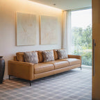 A modern living room with a tan leather sofa, patterned pillows, and the Blush Sketch Set of 2 art prints on the wall. A decorative vase sits nearby, while sheer curtains let light stream in through a large window overlooking greenery.