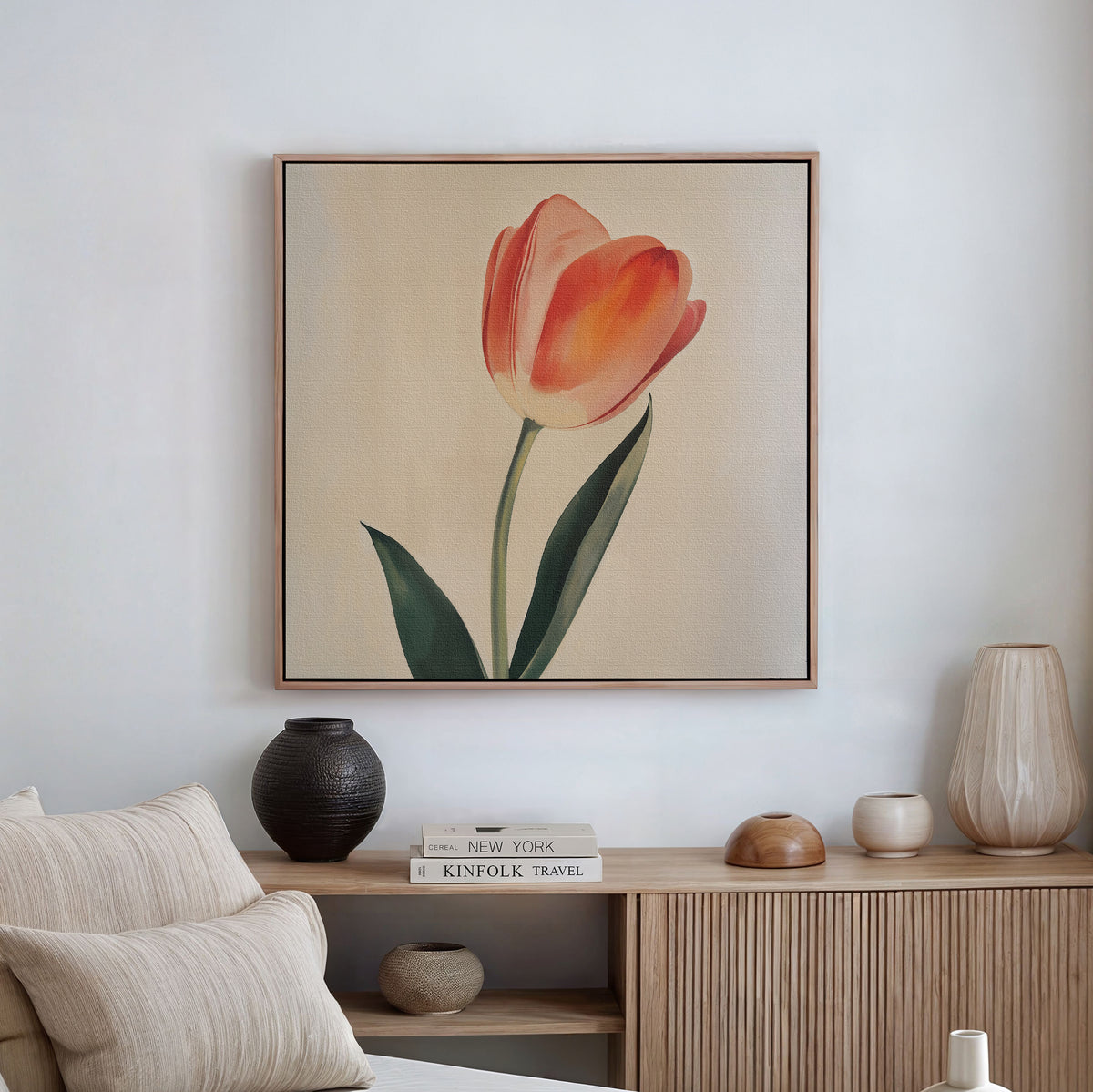 The Blush Tulip Canvas Art, showcasing an orange tulip with green leaves, is displayed above a wooden sideboard with vases and books. A beige sofa with striped cushions completes this modern minimalist decor.