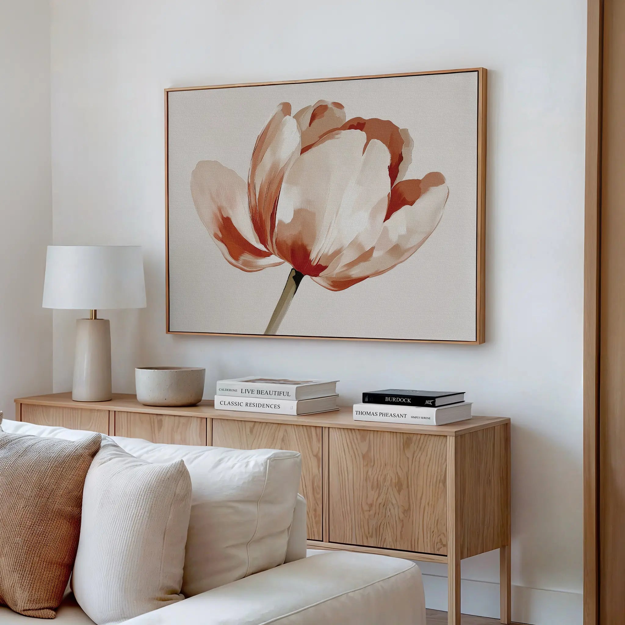 A modern living room with a light wood sideboard, white table lamp, decorative bowls, stacked books, and striking Blush Tulip Canvas Art featuring red and beige flowers above a cream-colored sofa.