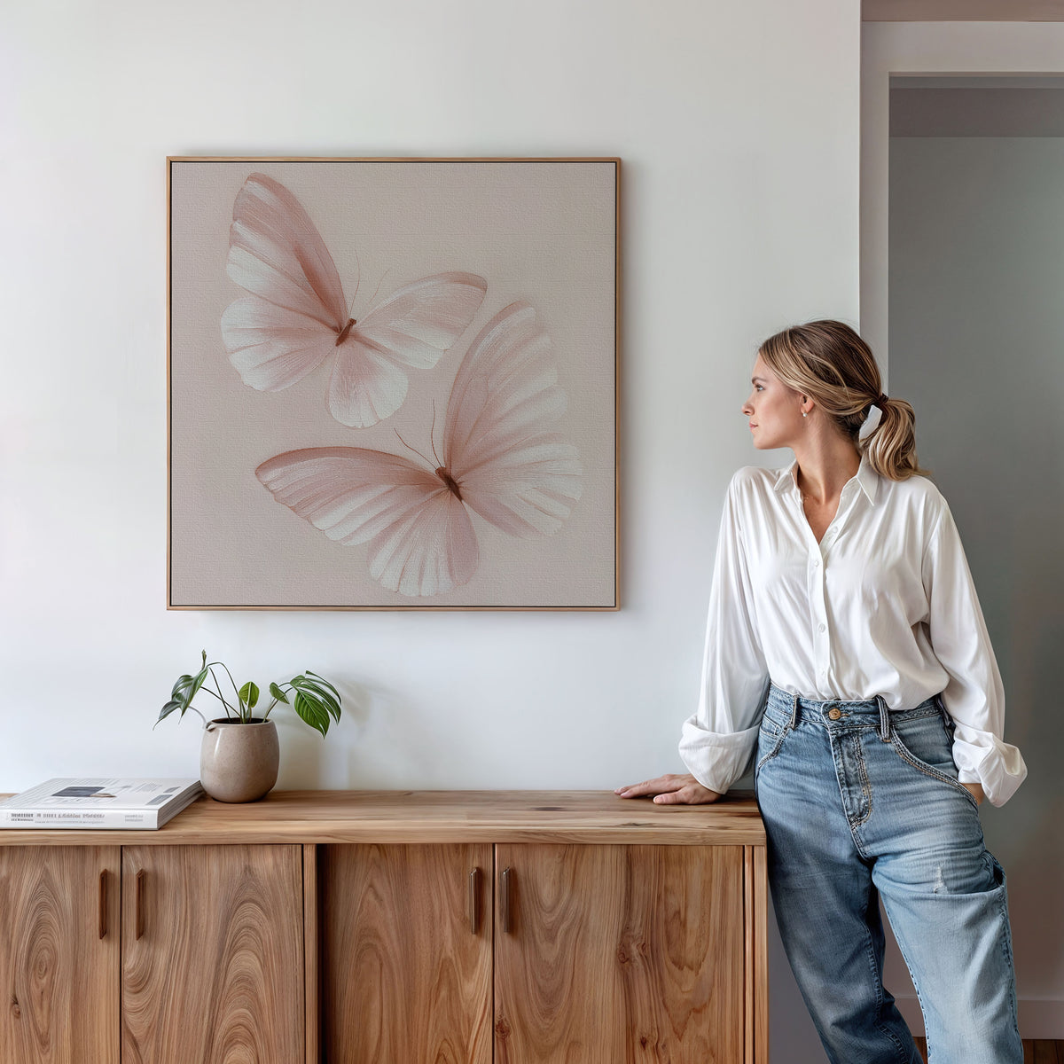 A woman in a white blouse and jeans stands by a wooden cabinet, admiring the Blush Wings Canvas Art with two light pink butterflies. A potted plant and book enhance the blush pink decor on the white wall.